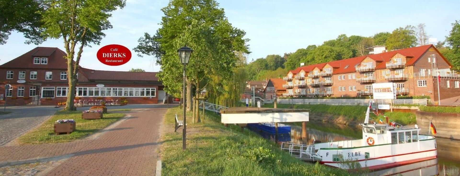 The image features a scenic view of a waterfront hotel with a boat docked beside it. Lush green trees line the pathway leading to the hotel, which has a red sign. The setting conveys a tranquil atmosphere near the water.