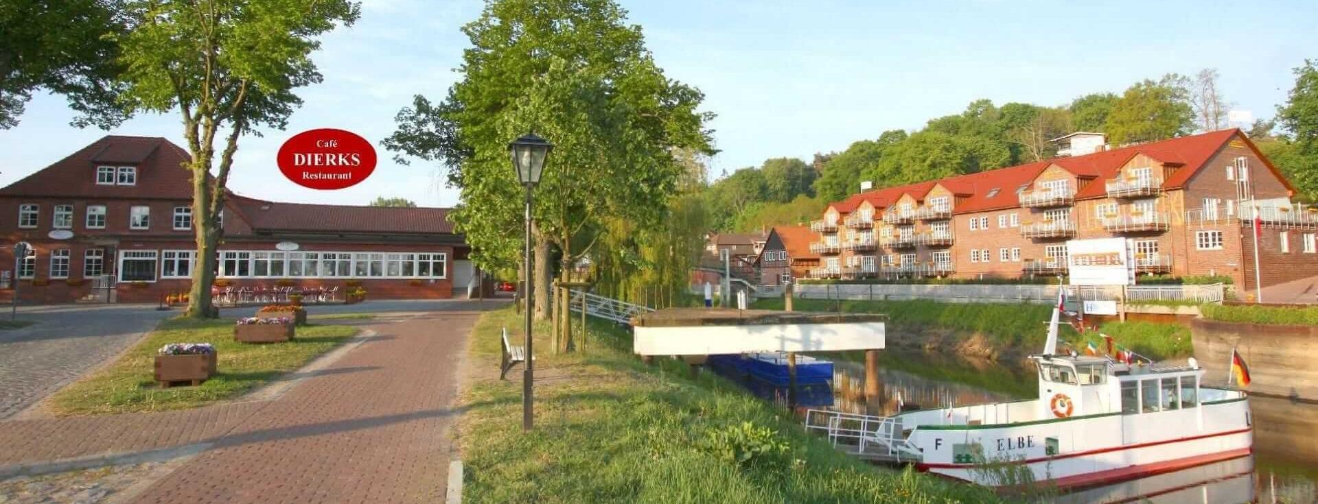 Das Bild zeigt den Hafen von Hitzacker mit einem Hotel im Hintergrund. Auf der linken Seite ist ein markantes Gebäude zu sehen, während rechts eine Flusslandschaft mit einem kleinen Boot zu erkennen ist. Grünflächen und Bäume umrahmen die Szene.